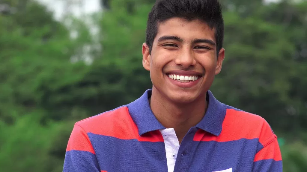 Hispanic teen boy smiles and laughs for the camera in a outdoors and forest-like area. 